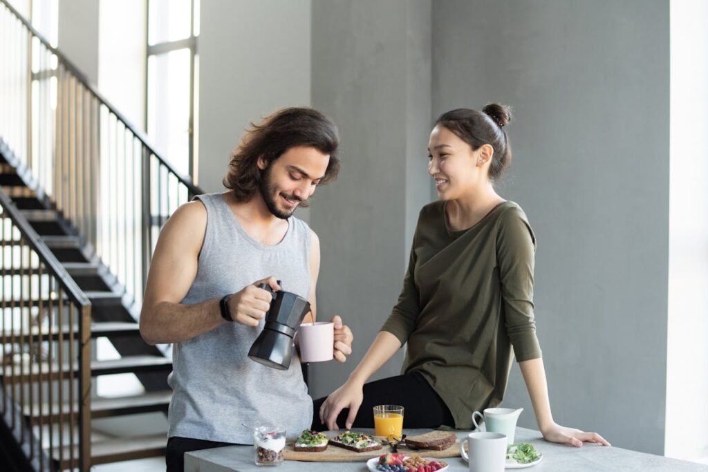 A couple enjoy a casual breakfast indoors with coffee and food, smiling and relaxed.